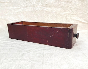 May include: A rectangular wooden drawer with a dark reddish-brown finish. The drawer has a small, round, black knob on one end. The interior of the drawer is a lighter brown. The drawer sits on a white, textured surface.