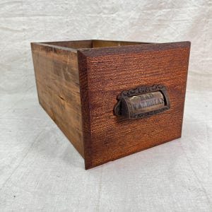 May include: A wooden drawer with a dark brown front and a lighter brown body. The drawer has a metal handle with the words "BAD VALERIAN" on it. The drawer is likely an antique or vintage piece.
