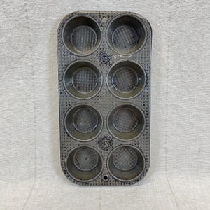 May include: Vintage metal muffin tin with eight round cups. The tin has a rectangular shape with a textured surface and small holes around the edges. The cups have a diamond pattern on the bottom. The words "OVENES" and "USA" are stamped on the tin.