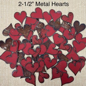 May include: A pile of 2-1/2 inch metal hearts in red and brown with a rustic finish. The hearts are scattered on a beige fabric background. 2-1/2" Metal Hearts