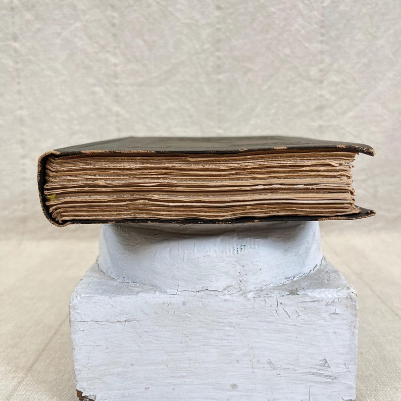 May include: A side view of an antique book with a dark cover and aged, yellowed pages. The book is resting on a white, weathered, pedestal-like structure, suggesting a vintage or historical theme. The book's spine is visible, and the pages show signs of age.