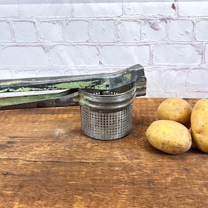 May include: An antique potato ricer with green handles and a perforated metal base, next to three raw potatoes on a wooden surface. The ricer shows signs of wear, suggesting its age and use.