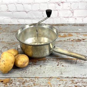 May include: A vintage, silver-toned food mill with a black handle, alongside three yellow potatoes. The mill has a bowl-shaped design with a handle and a rotating mechanism. The potatoes are of varying sizes.