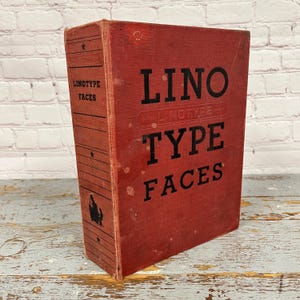 May include: A vintage red book titled "Linotype Faces" with black lettering. The book has a worn appearance, with visible wear and tear on the cover. The spine features the text "Linotype Faces" vertically. The book is likely a reference guide for typography.