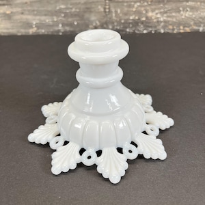 May include: A white, vintage-style candle holder. The base features a decorative, openwork design with leaf-like shapes. The candle holder has a round top and a ribbed, bulbous body. The overall design is ornate and elegant.