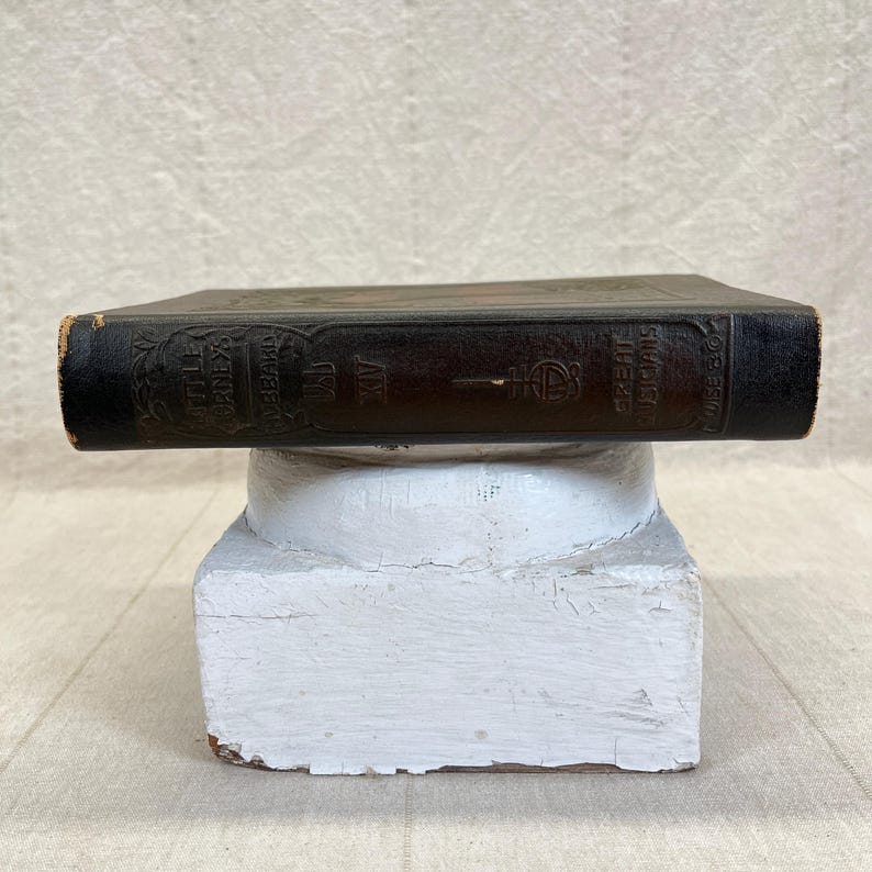 May include: A vintage, dark brown book titled "The Great Musicians" is displayed on a white, distressed pedestal. The book's cover features embossed lettering and decorative elements. The book is a classic piece of literature.