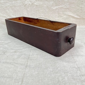 May include: A rectangular, dark brown wooden box with a rounded knob on one end. The interior of the box is a lighter brown color. The box appears to be vintage or antique, with visible wear and tear.