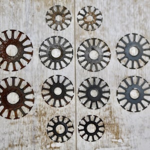 May include: Arrangement of various sizes of circular metal objects with a radial spoke design. The metal has a dark, aged appearance, with some showing signs of rust. The objects are arranged on a weathered, white wooden surface.