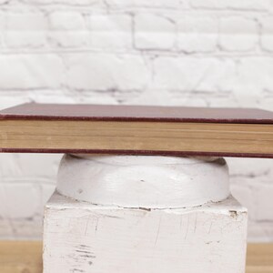 Pu&ograve; includere: Un libro chiuso con copertina rigida, di colore marrone con dorso dorato, &egrave; appoggiato su un piedistallo bianco. Il piedistallo &egrave; su una superficie di legno verniciata di bianco.