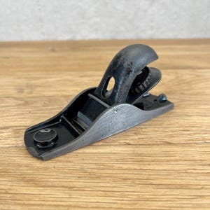 May include: A small, vintage hand plane with a black body and a gray blade. The plane has a curved handle and a metal lever cap. It is designed for woodworking and smoothing surfaces. The tool is made of metal and has a worn appearance.
