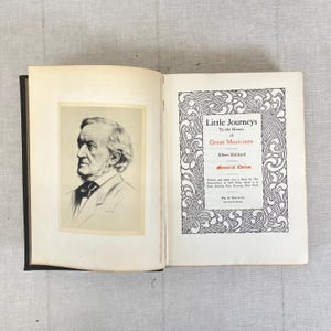 May include: An open book with a black spine, featuring a portrait of a man on the left page and decorative text on the right. The text on the right page reads "Little Journeys to the Homes of Great Musicians" by Elbert Hubbard.