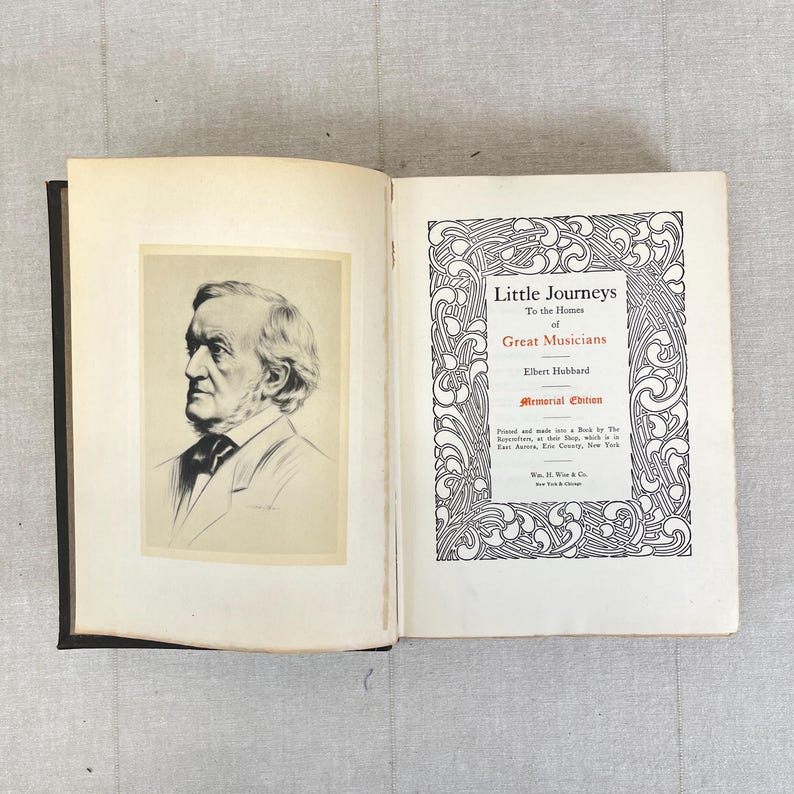 May include: An open book with a black spine, featuring a portrait of a man on the left page and decorative text on the right. The text on the right page reads "Little Journeys to the Homes of Great Musicians" by Elbert Hubbard.