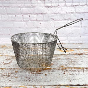 May include: A metal mesh fryer basket with a long handle. The basket is round and has a fine mesh pattern. The handle is attached to the side of the basket and curves upward. The basket is sitting on a weathered wooden surface.