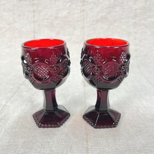 May include: Two ruby red vintage goblets with ornate, embossed designs. The glasses have a hexagonal base and a stem that leads to a detailed bowl. The interior of the glasses is a bright red. The goblets are likely made of glass.