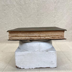May include: A vintage, leather-bound book with aged, yellowed pages. The book rests on a white, pedestal-like stand. The book's cover is dark, and the pages show signs of wear, suggesting age and use. The stand has a distressed, textured appearance.