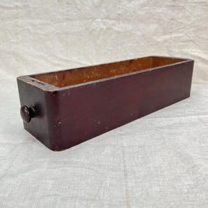 May include: A rectangular, dark burgundy wooden drawer with a round knob on the front. The drawer is open, revealing a lighter brown interior. The wood shows signs of age and wear, adding to its vintage appeal.