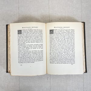 May include: Open book with text about Wolfgang Mozart. The pages are yellowed with age, and the text is in black. The book is open to pages 312 and 313, with text on both pages. The book is on a light gray surface.