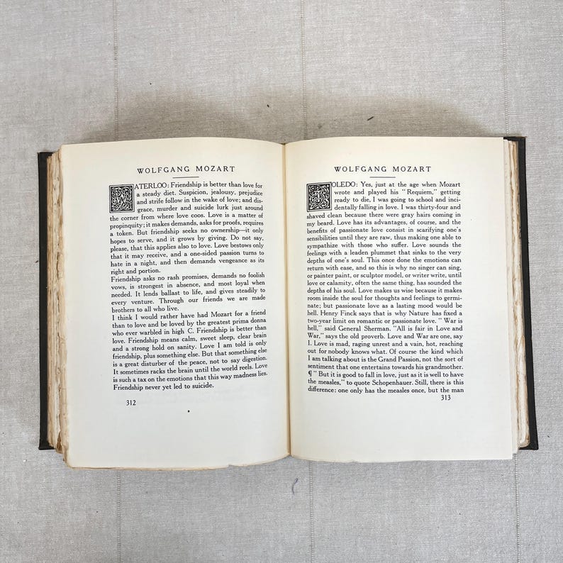 May include: Open book with text about Wolfgang Mozart. The pages are yellowed with age, and the text is in black. The book is open to pages 312 and 313, with text on both pages. The book is on a light gray surface.