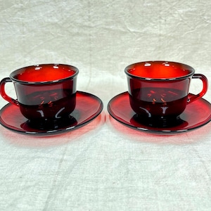 May include: Two matching ruby red glass teacups and saucers. The cups have a rounded shape with a handle, and the saucers are oval. The set is made of transparent glass, allowing light to pass through. The color is a deep, rich red.