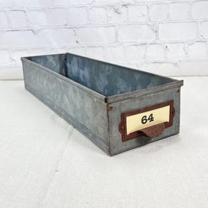 May include: A rectangular galvanized metal storage bin with a label holder. The label holder has the number 64 printed on a cream-colored label. The bin is made of corrugated metal and has a rustic, industrial aesthetic.