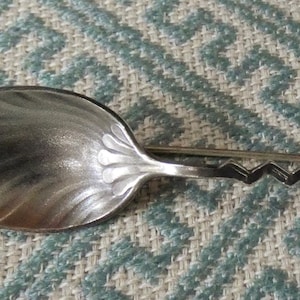 May include: A silver spoon with a decorative handle. The spoon's bowl has a swirling pattern, and the handle features a zigzag design and a detailed, ornate end. The spoon is set against a patterned blue and white background.