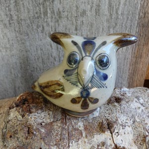 May include: A small ceramic owl figurine with brown, blue, and white painted details. The owl has large, round eyes and a detailed beak.