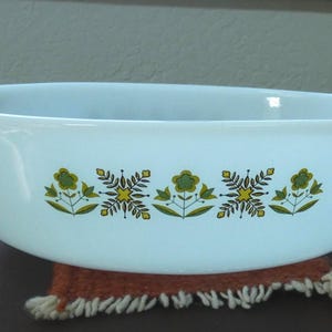 May include: White oval casserole dish with handles, decorated with a floral pattern in green, yellow, and brown. The dish is sitting on a brown and tan woven coaster. A vintage kitchenware item.