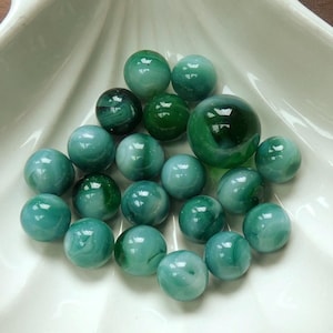 May include: A collection of round glass marbles in various shades of green, ranging from light mint to deep forest green. The marbles feature swirling patterns of opaque and translucent glass, with one larger marble positioned near the top right.