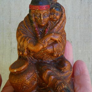 May include: A carved figurine depicting a Native American woman holding a child. The woman wears a red headband and traditional clothing. The figurine is brown with dark accents and has a detailed, textured surface.