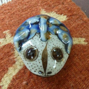 May include: A small, rounded ceramic object with a glossy finish, featuring a white base with brown speckles and a dark brown central marking. The top section is decorated with a vibrant blue and brown pattern, resembling stylized hair or a crown.