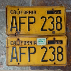 May include: Two vintage California license plates. The top plate is from 1956, and the bottom plate is from 1962. Both plates are yellow with black lettering and numbers. The plates read "CALIFORNIA" and "AFP 238".