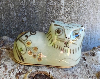 May include: A ceramic owl figurine in a seated position, painted in shades of pale green and brown. The owl has large, round eyes and decorative floral accents on its side. The figurine is resting on a textured wooden surface.