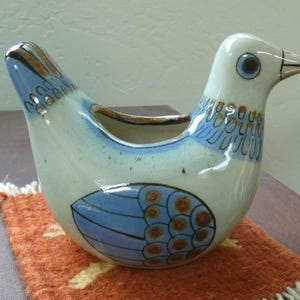May include: A ceramic bird-shaped container with a light blue and white glaze. The bird has blue and brown wing and tail details, and a dark eye. The container sits on a small orange woven mat.