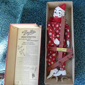 May include: A vintage Hazelle's marionette clown doll with a white face, red hat, and red outfit featuring white polka dots. The doll is inside a cardboard box with a wooden control bar and strings, alongside a box lid printed with operating instructions.