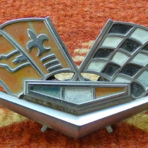 May include: A vintage car emblem with a silver base and colorful enamel details. The emblem features two flags, one with a checkered pattern and the other with a fleur-de-lis design, set against an orange background. The emblem is displayed on an orange textured surface.