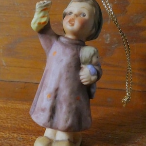May include: A ceramic figurine of a young girl in a mauve dress with orange polka dots, holding a doll and a yellow and orange object. The figurine has a gold string for hanging. The girl has rosy cheeks and is looking upwards.