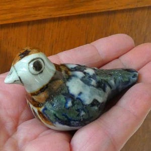 May include: A ceramic bird figurine with a white head, brown and black markings, and a body with blue and green patterns. The bird has a small white beak and a large eye. The figurine is held in a hand.