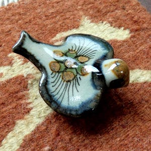 May include: A ceramic whistle with a light blue base, adorned with brown and green floral designs and black accents. The whistle has a rounded body and a spout, resting on a textured, reddish-brown surface.
