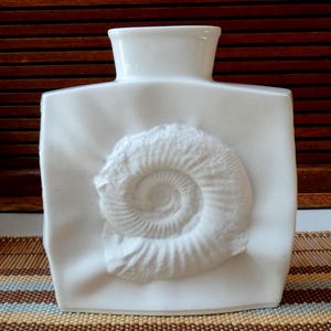 May include: A white ceramic vase with a square body and a rectangular opening. The front of the vase features a raised, detailed relief of an ammonite fossil. The vase is a neutral colour and has a smooth finish.