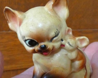 May include: A vintage ceramic figurine of a cat and mouse. The cat is light beige with brown accents, black eyes, and a red tongue. The mouse is light beige with brown accents. The cat is winking and the mouse is hugging the cat.