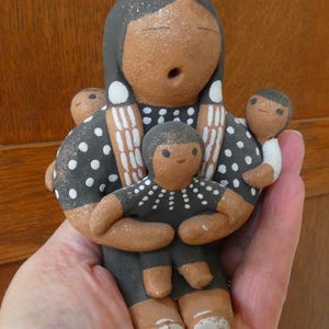 May include: A hand-painted clay figurine featuring a central adult figure with a dark gray head and torso, wearing a black garment with white polka dots. The figure holds a smaller child in front, with two additional small children attached to the sides.