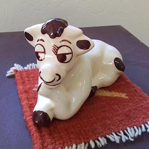 May include: A glossy white ceramic cow figurine in a reclining position, featuring dark brown painted spots on its head, ears, and body, with dark brown painted eyes, eyelashes, and a snout, resting on a small, square, textured orange rug.