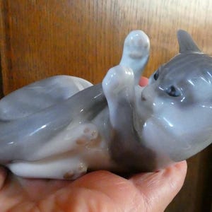 May include: A ceramic figurine of a playful cat, lying on its back with paws raised. The cat is primarily gray with white accents and blue eyes. The figurine is a decorative item.
