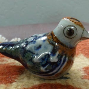 May include: A small ceramic bird figurine featuring a glossy glaze with a color palette of white, deep blue, and earthy brown. The bird has a rounded body, a pointed beak, and a painted circular eye, with a smooth, reflective surface finish.