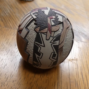 May include: A round, ceramic ornament with a geometric design in black, white, brown, and red. The design features a stylised figure with a feathered headdress.