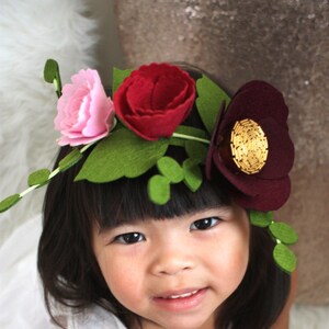 May include: A floral crown featuring felt flowers in shades of light pink, deep red, and dark burgundy. The arrangement includes green felt leaves and stems, with the dark burgundy flower accented by a circular center of gold metallic beads.