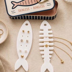 May include: A rectangular tin box labeled not SARDINES SKINLESS + BONELESS with a pull tab, two white fish-shaped jewelry organizers, and a small ceramic dish containing gold rings. One organizer holds stud earrings, the other holds gold chains.