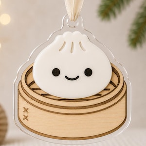 May include: A hanging ornament featuring a white dumpling with a simple black face sitting inside a light-coloured wooden bamboo steamer basket. The design is encased in a clear acrylic shape and includes a cream-coloured ribbon for hanging purposes.