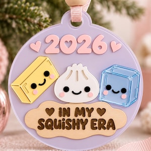 Op de afbeelding: Een ronde lavendelkleurige hanger met het jaartal 2026 in roze, voorzien van drie schattige figuren: een geel boterblokje, een witte dumpling en een blauw ijsblokje. Daaronder staat de tekst IN MY SQUISHY ERA op een licht houten wolk.