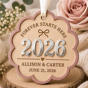 Op de afbeelding: Een geschulpt houten ornament met een oudroze rand en een wit lint. De tekst luidt FOREVER STARTS HERE, 2026, ALLISON & CARTER en JUNE 21, 2026. Het bevat een lasergegraveerde strik, een klein hartje en verhoogde zilveren 2026-cijfers.